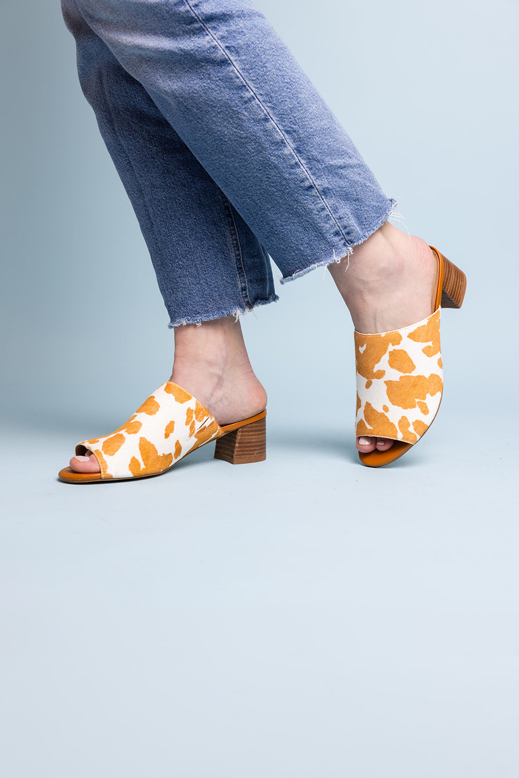 Tan and white cow print mule shoes for women, featuring a genuine calf-hair upper, synthetic lining, padded insole, and internal elastic gore for comfort. Designed with a low stacked leather heel, these slip-on mules are perfect for pairing with jeans, jumpsuits, or other outfits to add a playful, bold touch.