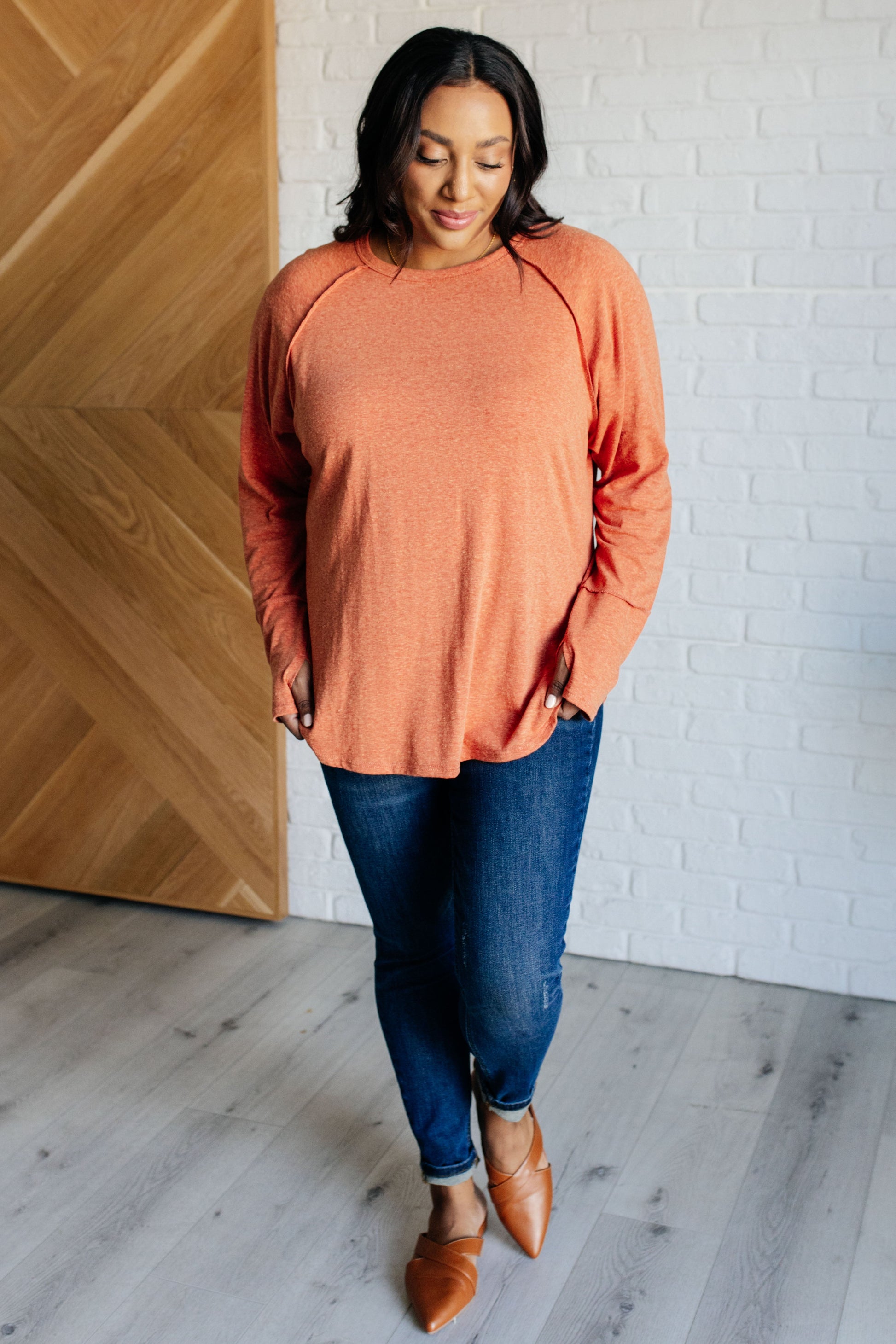 Pumpkin-colored long sleeve top with raglan sleeves, exposed seams, thumb hole cuffs, and scooped hemline.