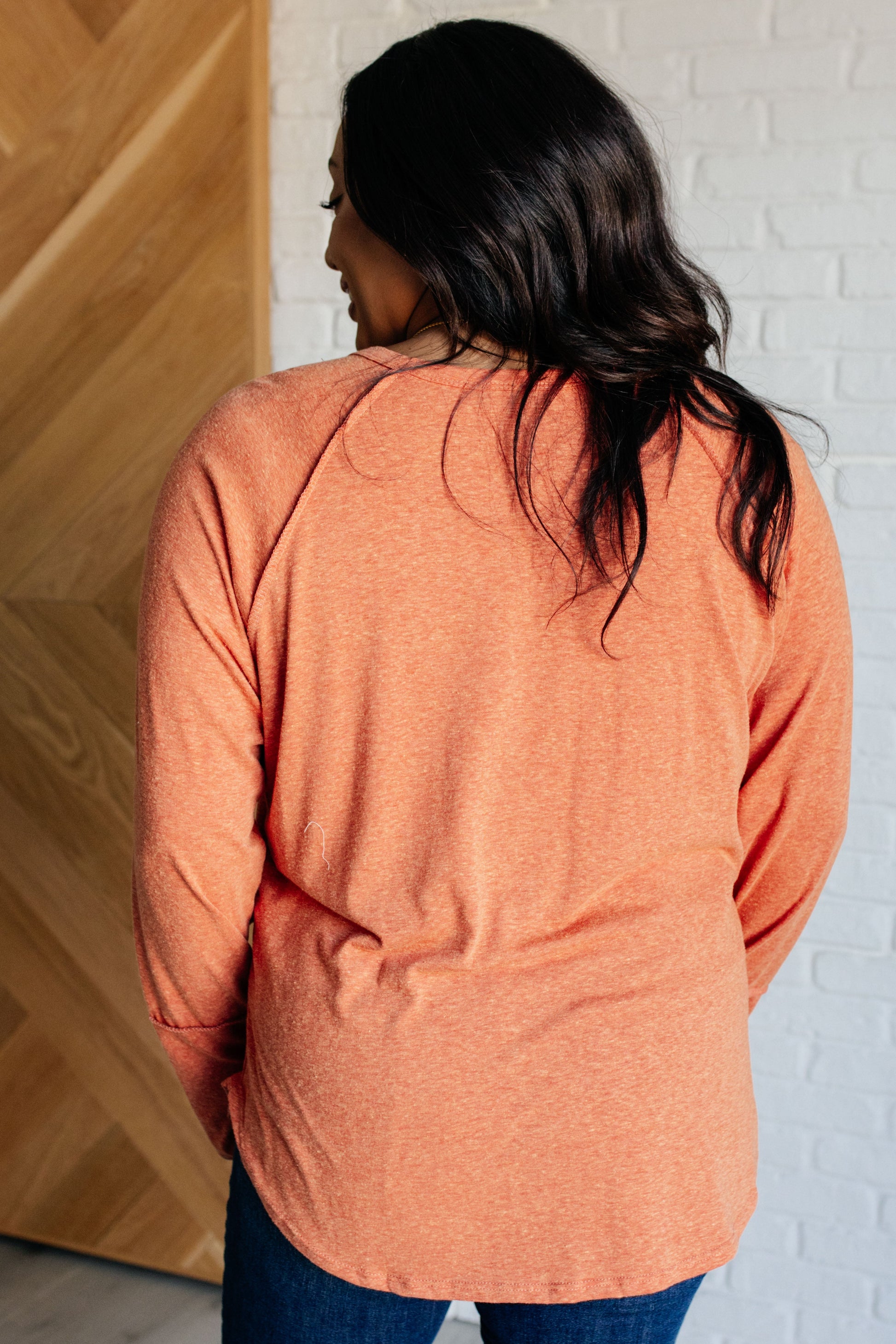 Pumpkin-colored long sleeve top with raglan sleeves, exposed seams, thumb hole cuffs, and scooped hemline.