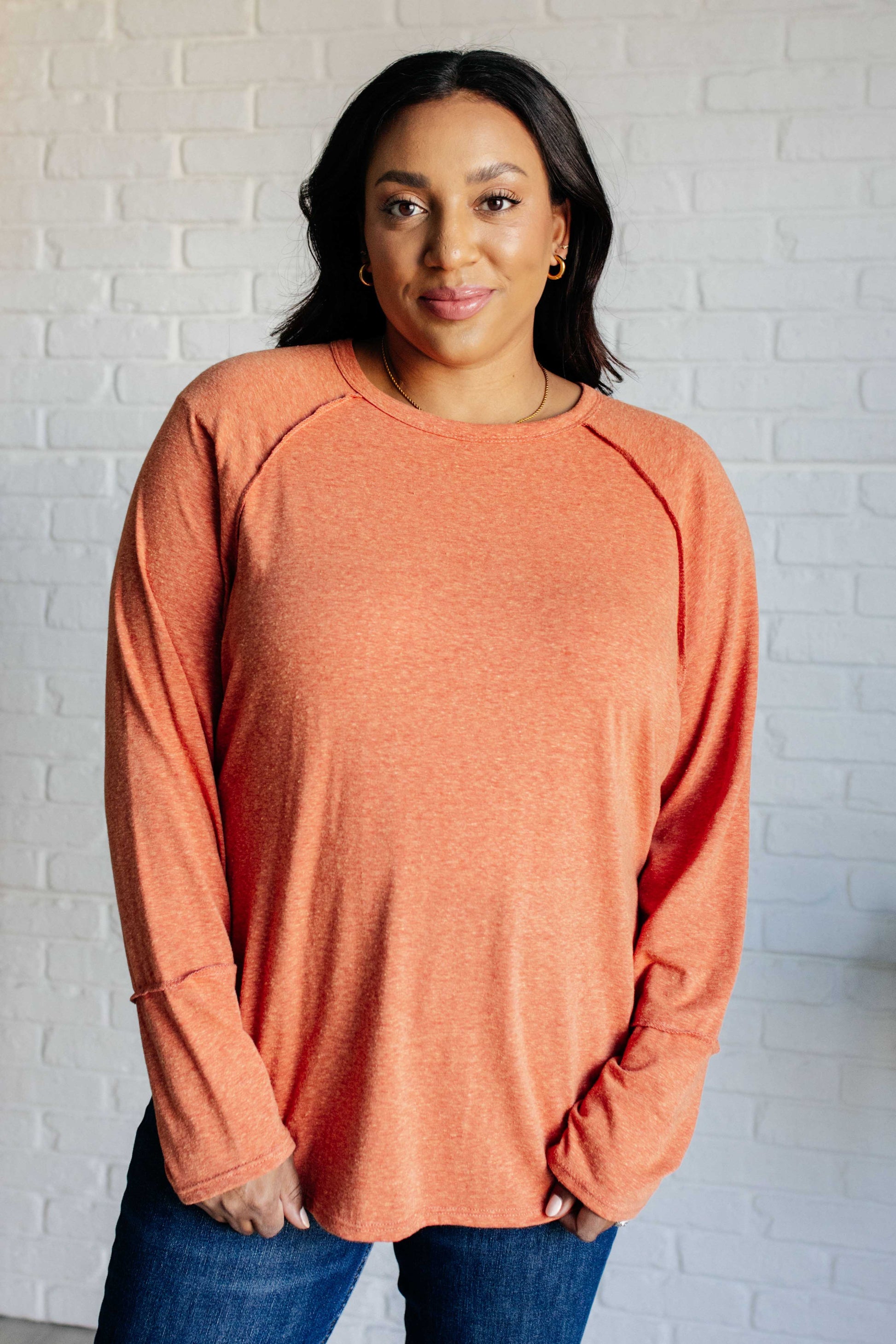 Pumpkin-colored long sleeve top with raglan sleeves, exposed seams, thumb hole cuffs, and scooped hemline.