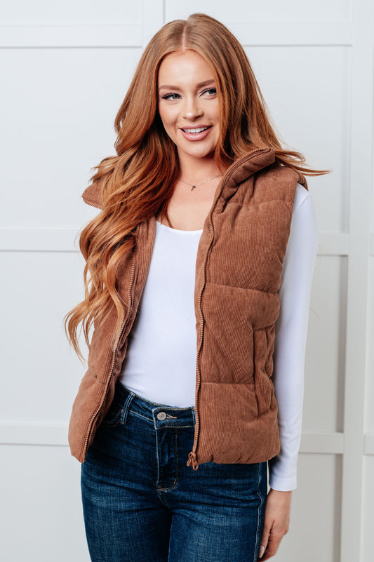 Brown corduroy puffer vest with a high-collared neckline, sleeveless design, full front zipper closure, functional welt pockets, and fully lined interior.