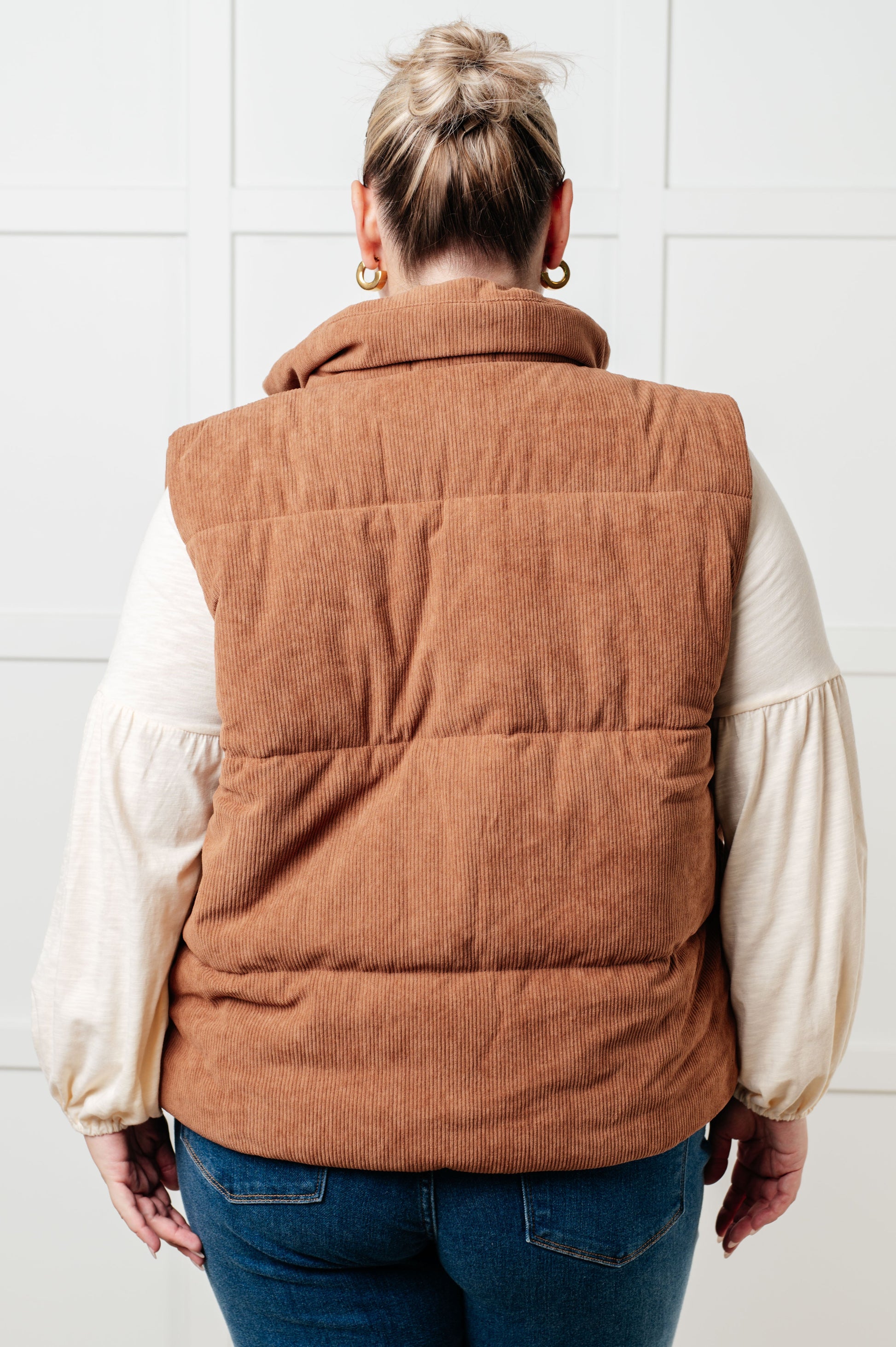 Brown corduroy puffer vest with a high-collared neckline, sleeveless design, full front zipper closure, functional welt pockets, and fully lined interior.