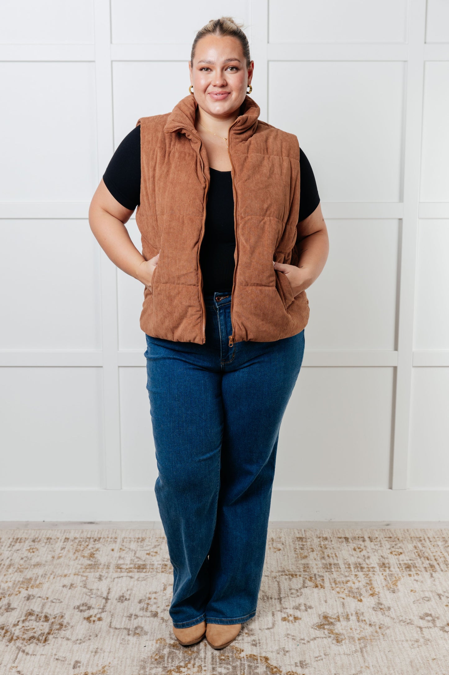 Brown corduroy puffer vest with a high-collared neckline, sleeveless design, full front zipper closure, functional welt pockets, and fully lined interior.