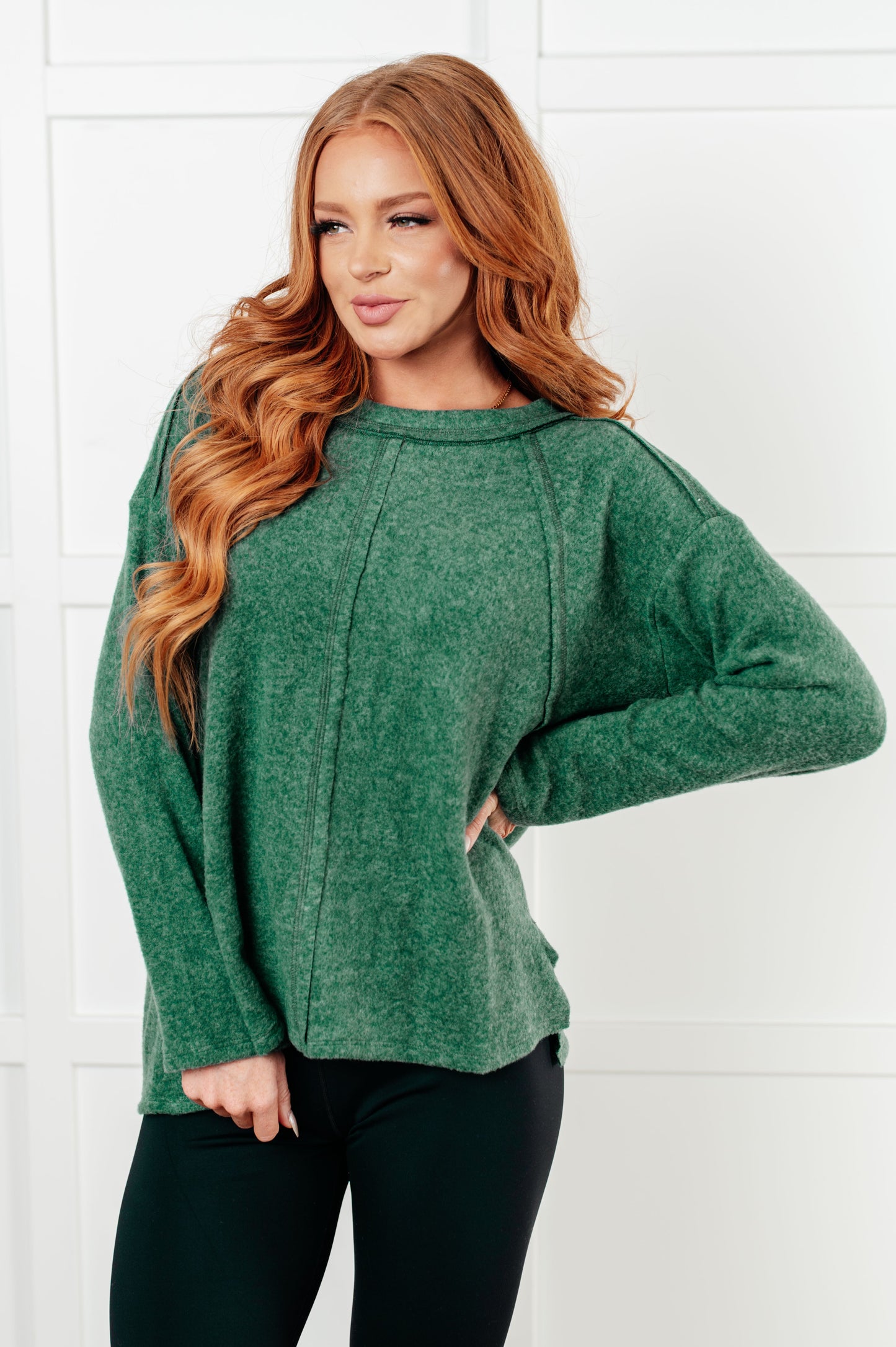 Women's Brushed Hacci Sweater in dark green featuring a round neckline, drop shoulders, stepped hemline, side slits, and raw seams. 