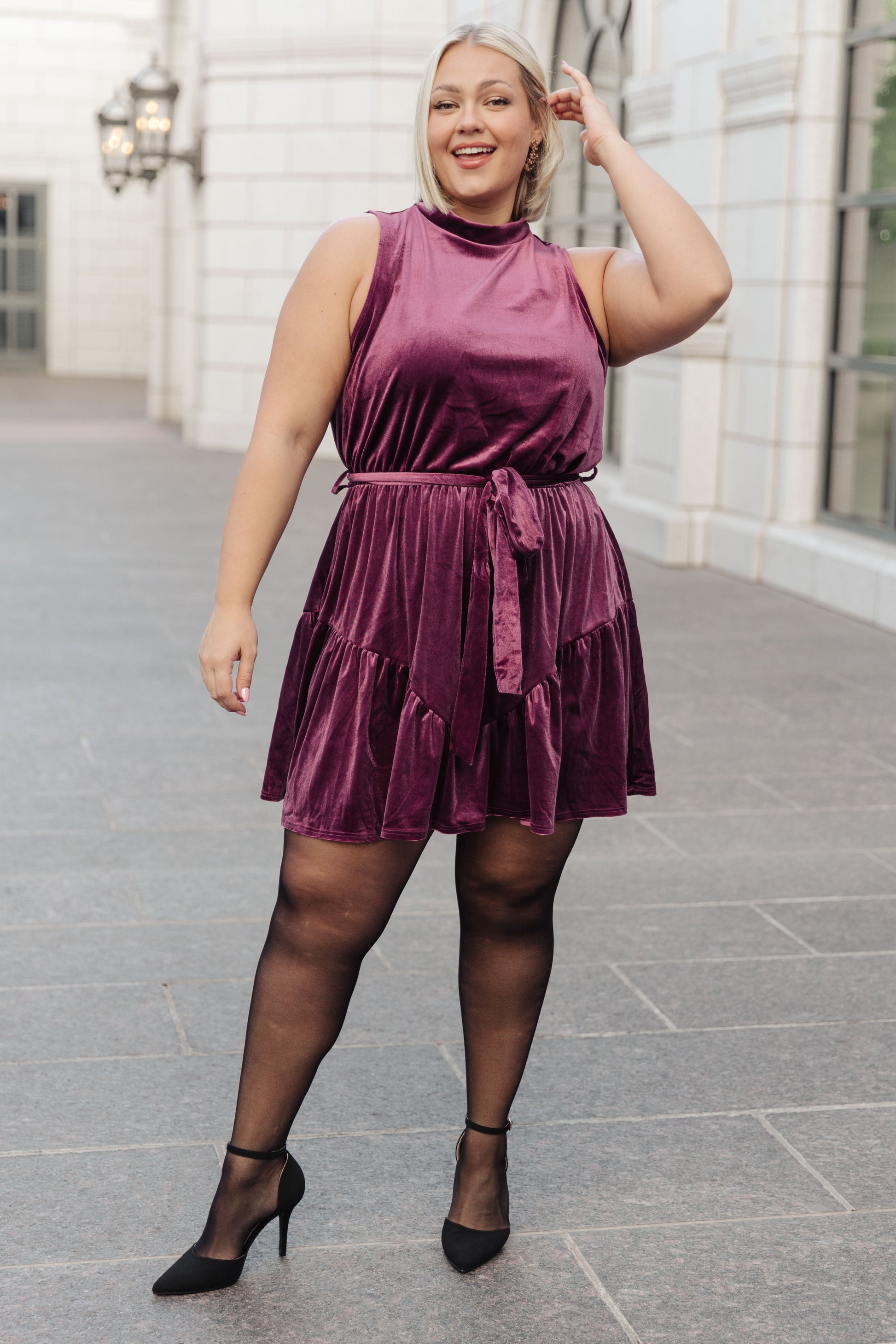 Purple velvet sleeveless dress with a mock neckline, keyhole button closure, and cased elastic waistband. Features a removable self-tie belt, ruffled hem, and is fully lined.