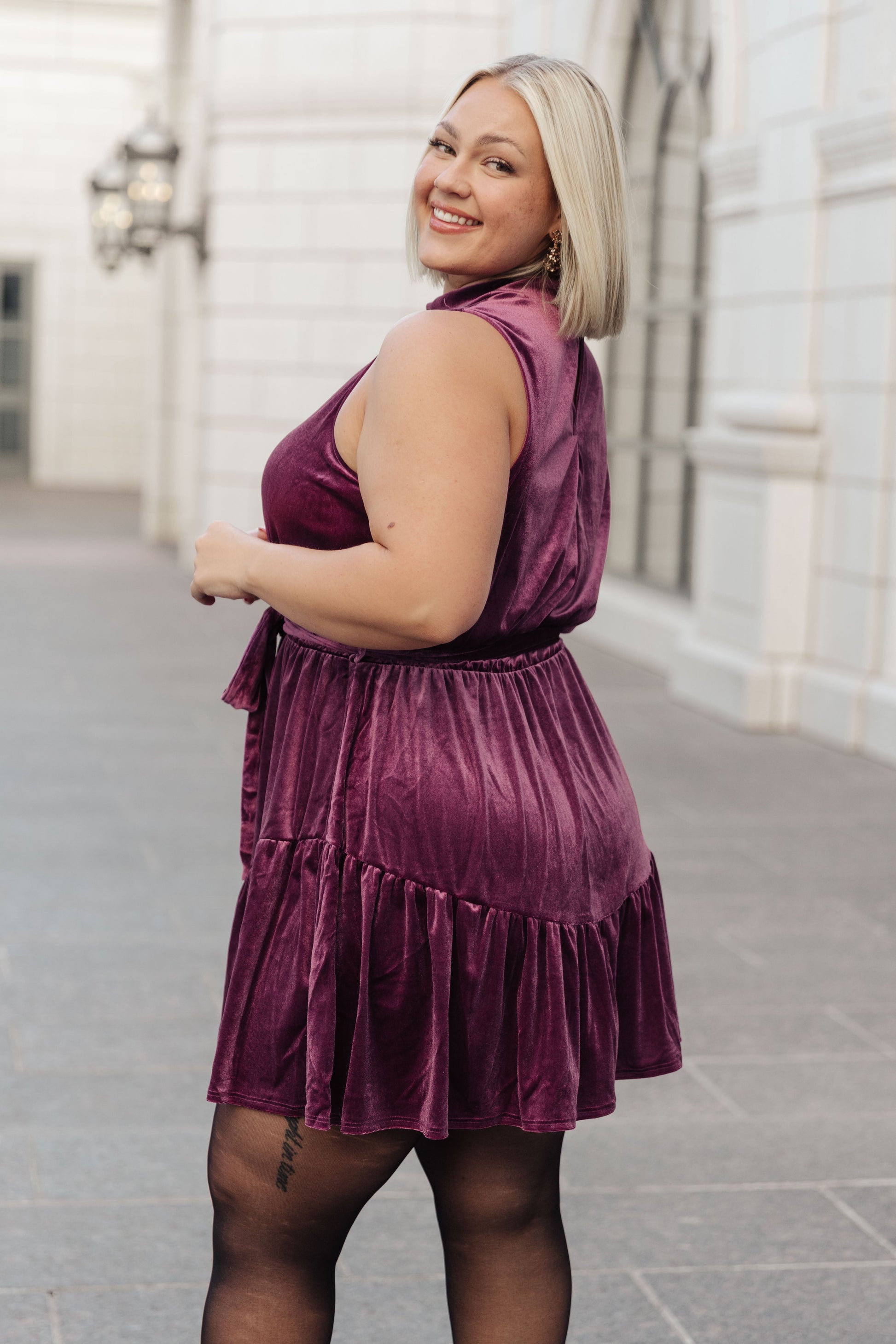 Purple velvet sleeveless dress with a mock neckline, keyhole button closure, and cased elastic waistband. Features a removable self-tie belt, ruffled hem, and is fully lined.