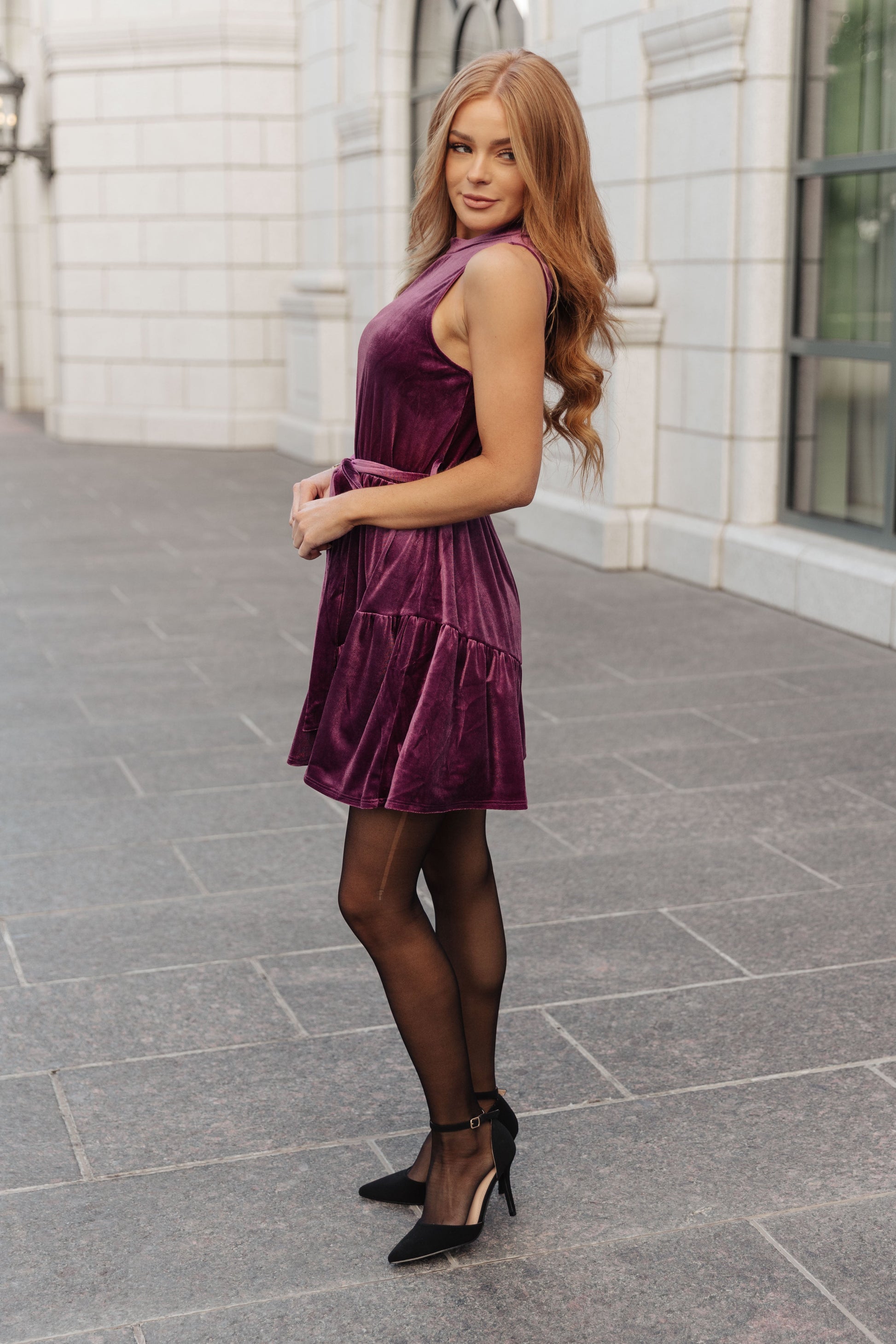 Purple velvet sleeveless dress with a mock neckline, keyhole button closure, and cased elastic waistband. Features a removable self-tie belt, ruffled hem, and is fully lined.