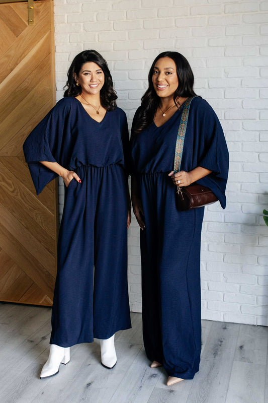 Navy crinkle woven wide-leg jumpsuit with V-neckline, kimono half sleeves, and elastic waistline. Features functional side pockets and keyhole button closure.