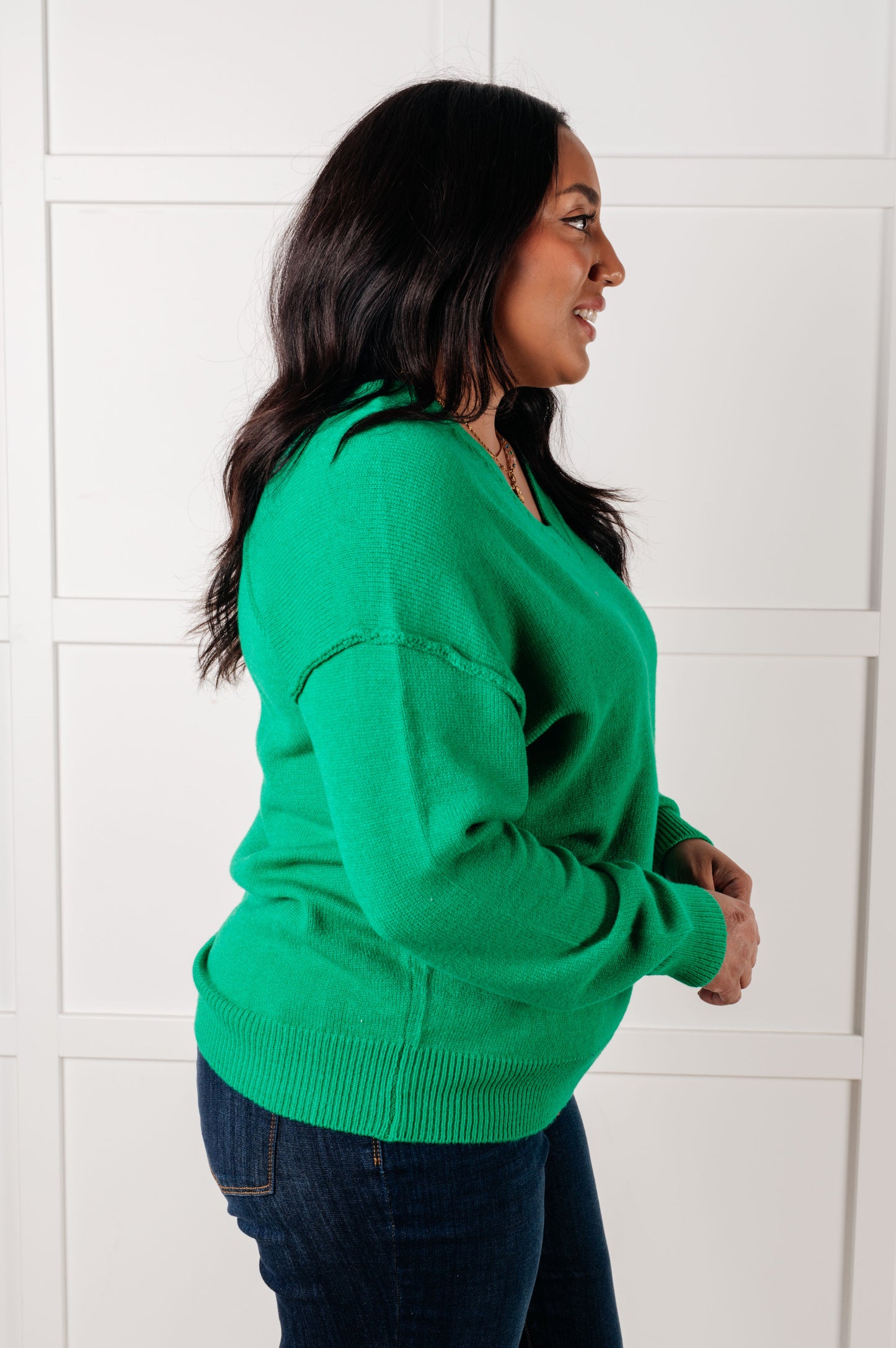 Side view of a plus size model wearing the Veru V-Neck Sweater in Green with ribbed cuffs and hem, drop shoulder design, exposed seams, and lightweight knit fabric, perfect for layering or standalone wear.