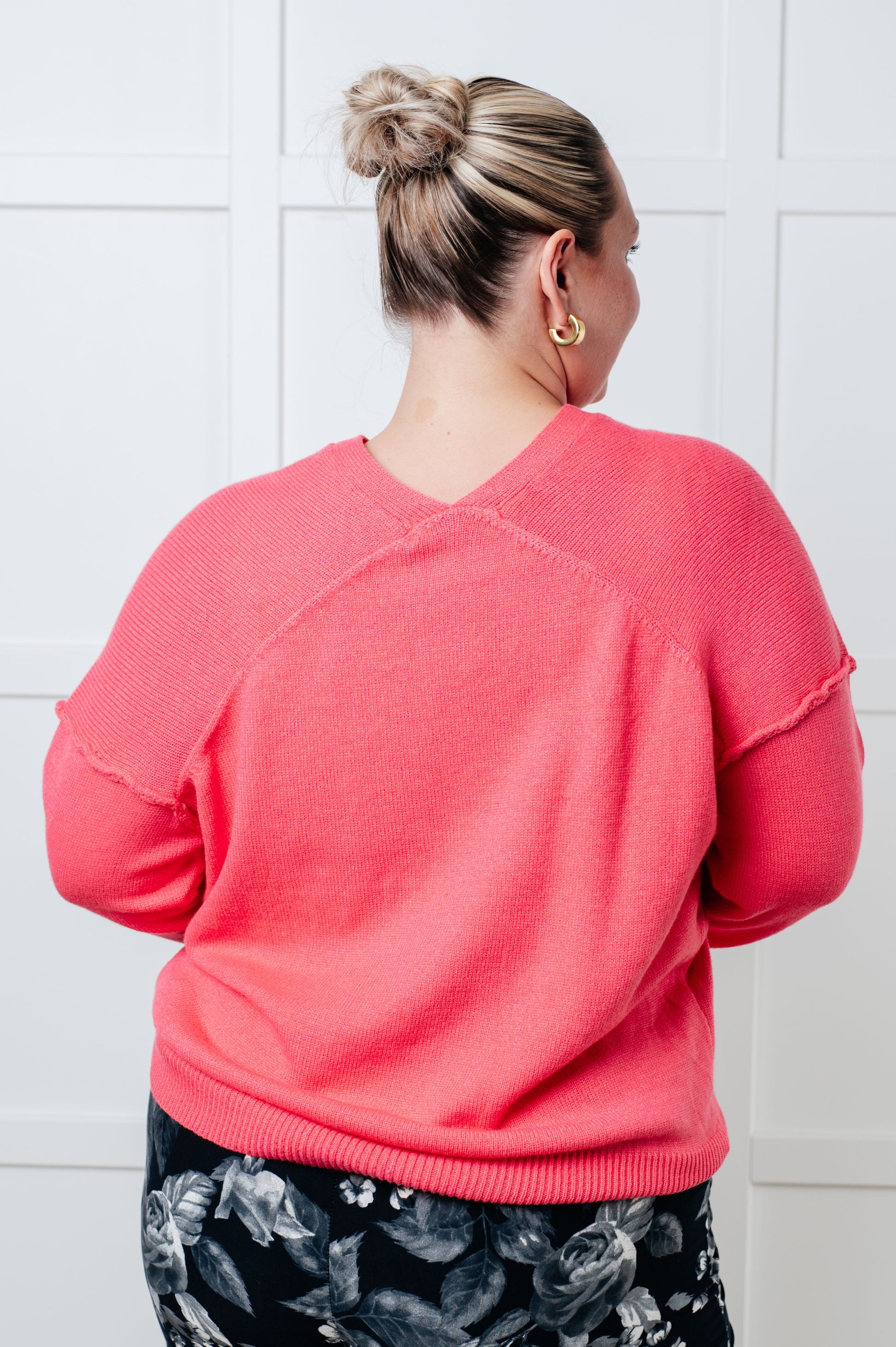 A women's lightweight sweater in pink, with a banded V-neckline, long drop-shoulder sleeves, a back V detail, and exposed seams. It has a banded ribbed hemline and sleeve cuffs.