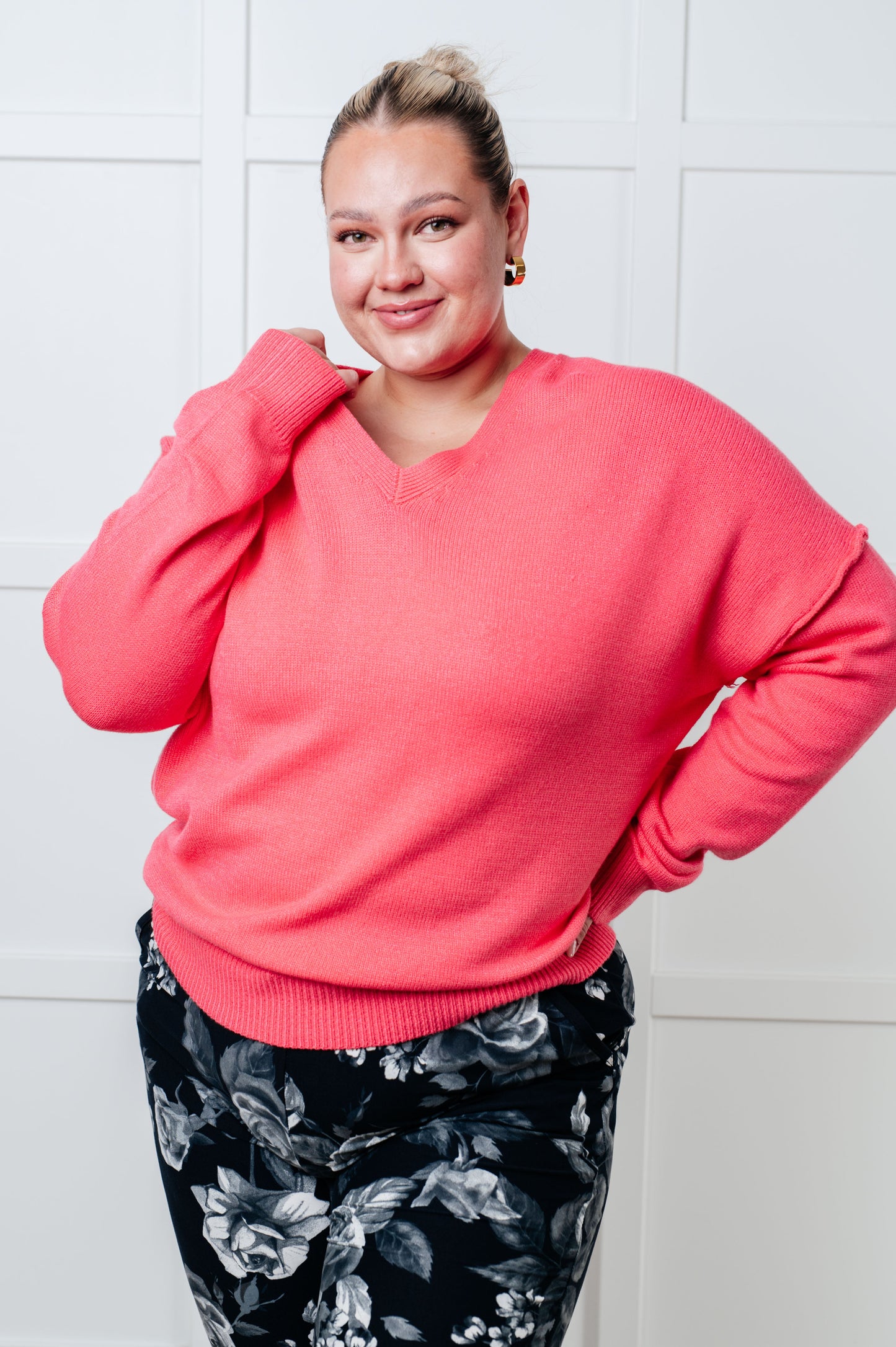 A women's lightweight sweater in pink, with a banded V-neckline, long drop-shoulder sleeves, a back V detail, and exposed seams. It has a banded ribbed hemline and sleeve cuffs.