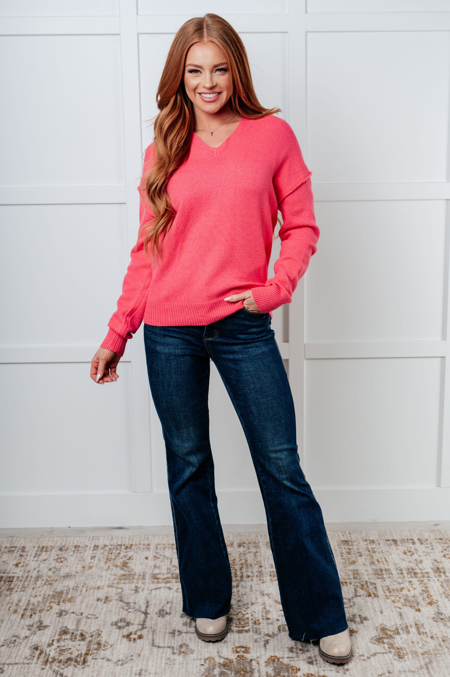 A women's lightweight sweater in pink, with a banded V-neckline, long drop-shoulder sleeves, a back V detail, and exposed seams. It has a banded ribbed hemline and sleeve cuffs.