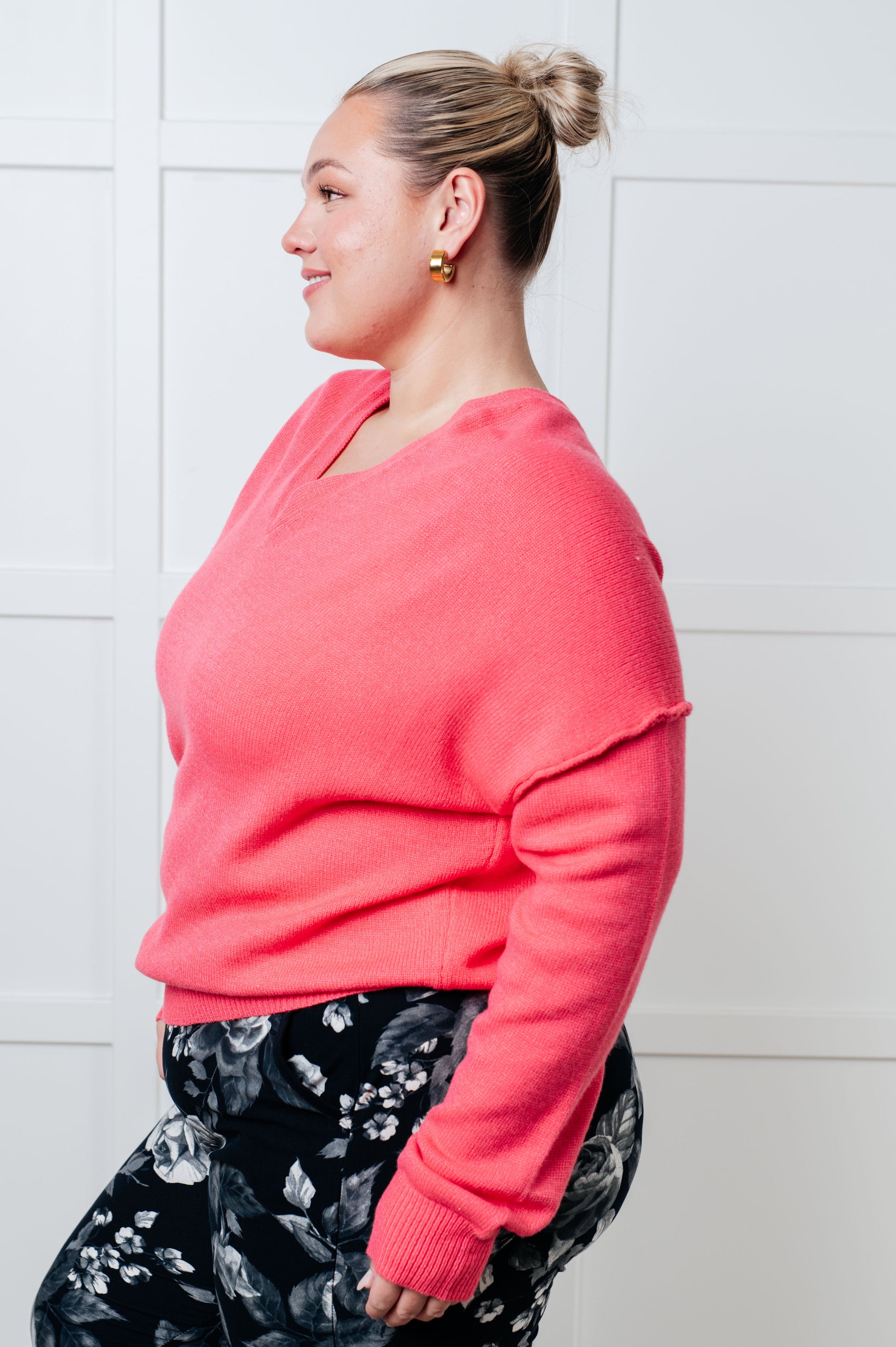 A women's lightweight sweater in pink, with a banded V-neckline, long drop-shoulder sleeves, a back V detail, and exposed seams. It has a banded ribbed hemline and sleeve cuffs.