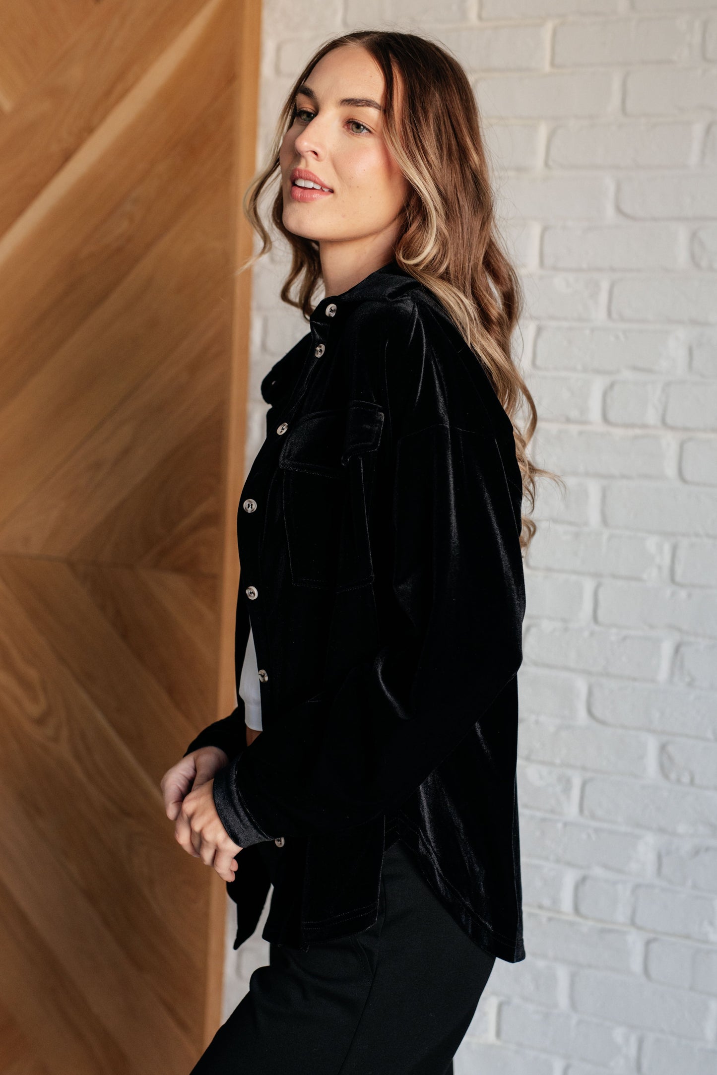 Black velvet knit button-down top with a collared neckline, long sleeves, and breast flap pockets. Features front button closure, buttoned sleeve cuffs, and a scooped hemline.