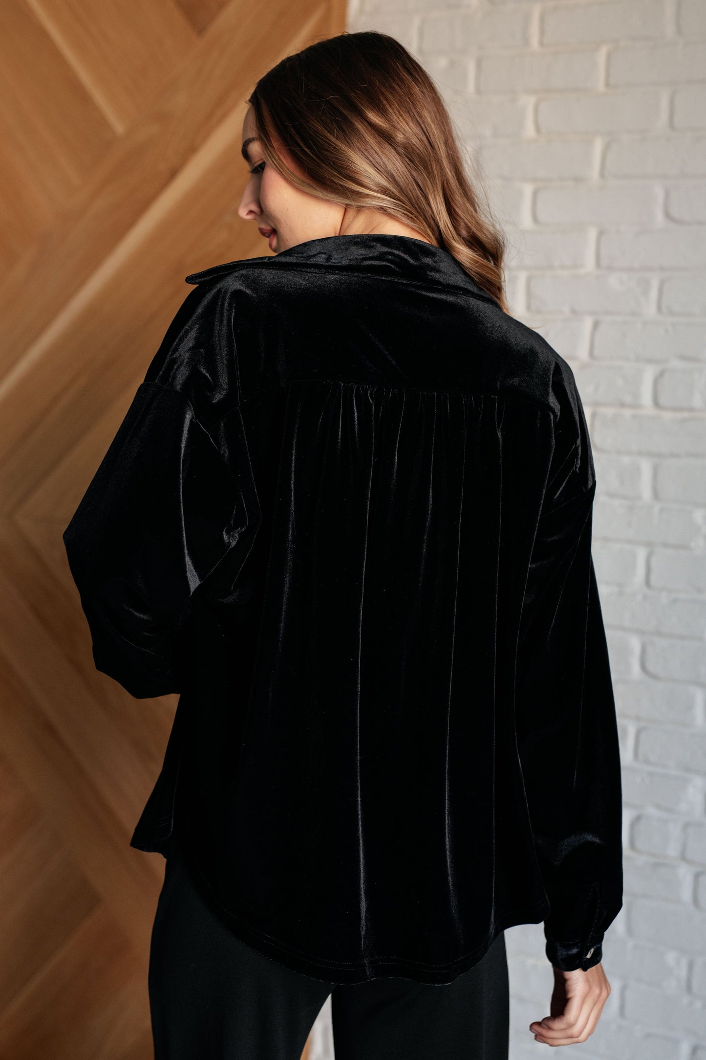 Black velvet knit button-down top with a collared neckline, long sleeves, and breast flap pockets. Features front button closure, buttoned sleeve cuffs, and a scooped hemline.