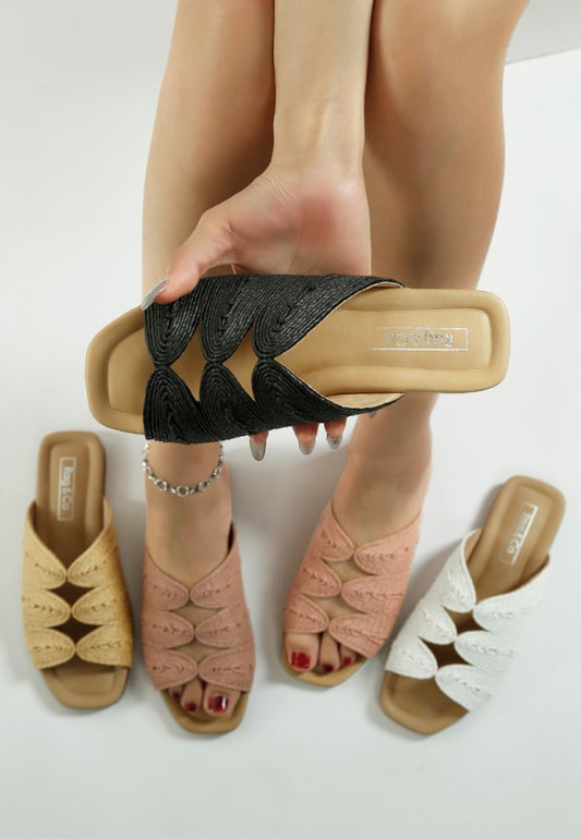 Woman wearing dusty pink raffia slip-on flats, with beige and white flats beside her, holding a black flat in her hand.