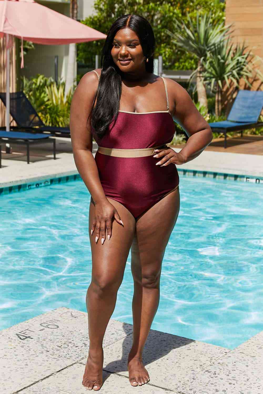 Wine-colored one-piece swimsuit with a contrast waistband, open back, shiny fabric, and a straight neckline.