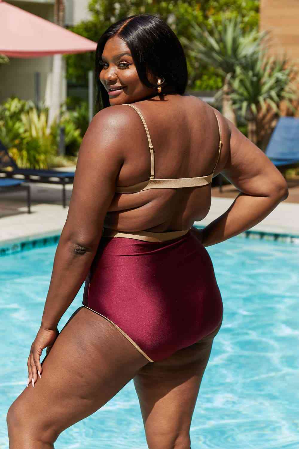 Wine-colored one-piece swimsuit with a contrast waistband, open back, shiny fabric, and a straight neckline.